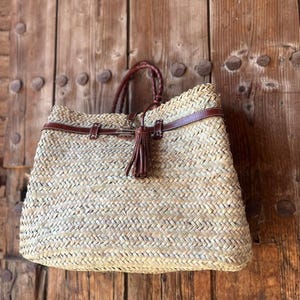 May include: Woven straw tote bag with brown leather detailing. It has a braided handle and a decorative tassel. The bag is rectangular and suitable for carrying various items.