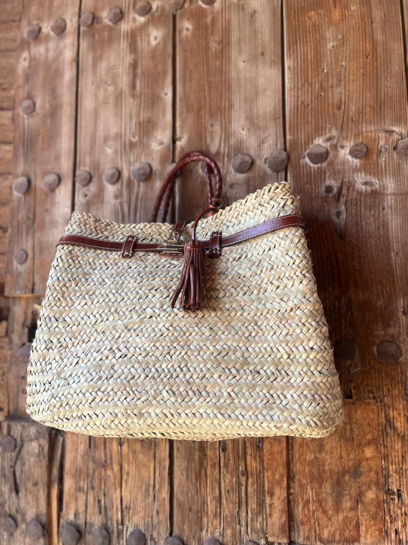 May include: Woven straw tote bag with brown leather detailing. It has a braided handle and a decorative tassel. The bag is rectangular and suitable for carrying various items.