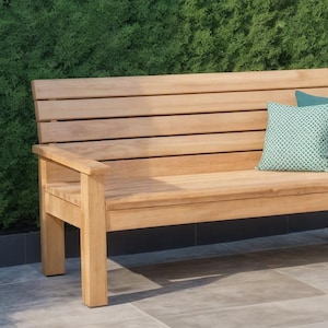 Op de afbeelding: Houten tuinbank met lattenrugleuning en armleuningen. Twee decoratieve kussens in teal en groen met patroon liggen op de bank. De bank is gemaakt van lichtgekleurd hout en staat tegen een achtergrond van groen gebladerte.