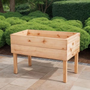 May include: A rectangular wooden planter box with four legs. The planter is made of light-colored wood and has a simple, clean design. The box is empty and ready for planting. The background features green bushes and a stone patio.