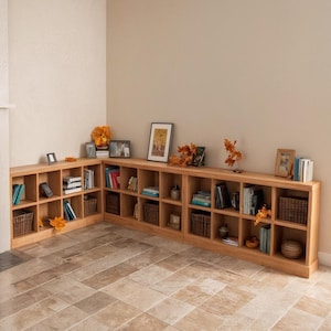 May include: A built-in, light-colored wooden bookcase with multiple cubbyhole shelves. The bookcase is filled with books, decorative items, and woven baskets. The bookcase is positioned in a corner of a room with a neutral-colored wall and a tiled floor.