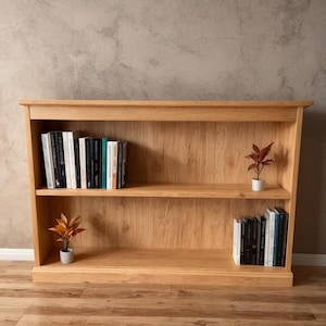 Puede incluir: Librería de madera clara de dos niveles. Los estantes están llenos de libros y pequeñas plantas en macetas. La librería tiene un diseño sencillo y clásico. Está hecha de madera maciza y es adecuada para una sala de estar o una oficina en casa. Mide unos 100 cm de ancho.