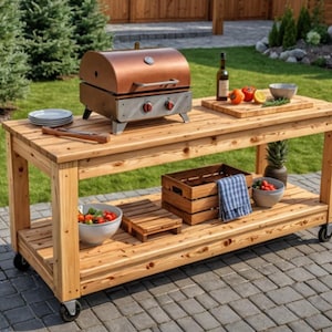 Könnte beinhalten: Ein rechteckiger Outdoor-Vorbereitungstisch aus Holz auf Rollen mit unterer Ablage, einem kupferfarbenen Tischgrill, einem Holzschneidebrett mit frischem Gemüse, einer Salatschüssel, einer Holzkiste, einem blau karierten Tuch und einer Ananas.