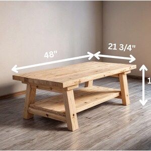 May include: A natural wood coffee table with a lower shelf. The table is 48 inches long, 21 3/4 inches wide, and 16 inches high. The table has a rectangular top and sturdy legs, showcasing a rustic design.