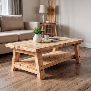 May include: A rustic, rectangular coffee table made of light-colored wood. The table has a lower shelf and sturdy, angled legs. A small white vase with greenery sits on top, alongside a book and a cup and saucer.