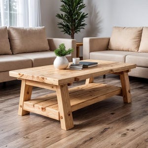 May include: A light-colored wooden coffee table with a lower shelf. The table has a rectangular top and sturdy legs. A small white vase with greenery, a cup, and books are on the table. The table is in a living room setting with beige sofas.