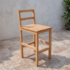 May include: A light-colored wooden bar stool with a rectangular seat and a backrest. The stool has four legs and a footrest. The wood grain is visible. The stool is outdoors, with plants and a wall in the background.