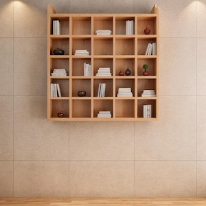 May include: A light brown, square, wall-mounted bookshelf with multiple cubby holes. The shelves are filled with stacks of white books and decorative items, including small vases and a miniature tree. The bookshelf is mounted on a beige tiled wall.