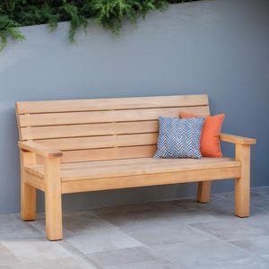 May include: A light brown wooden bench with a slatted back and armrests. Two decorative pillows, one orange and one blue with a chevron pattern, rest on the bench. The bench is outdoors against a gray wall.