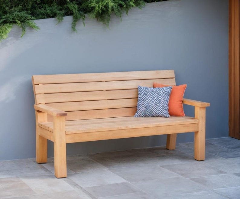 May include: A light brown wooden bench with a slatted back and armrests. Two decorative pillows, one orange and one blue with a chevron pattern, rest on the bench. The bench is outdoors against a gray wall.