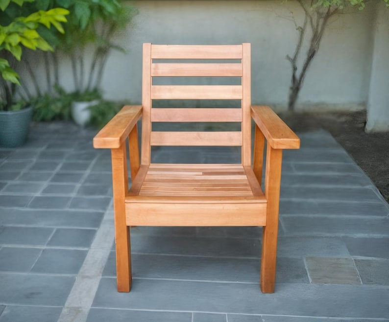 May include: A wooden outdoor chair with a natural finish. The chair has a slatted back and seat, with armrests. The chair is made of light brown wood and is placed on a grey tiled surface.