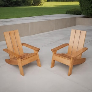 May include: Two wooden Adirondack chairs with a natural finish. The chairs have wide armrests and slatted backs. They are positioned on a concrete patio, with a grassy area and greenery in the background.