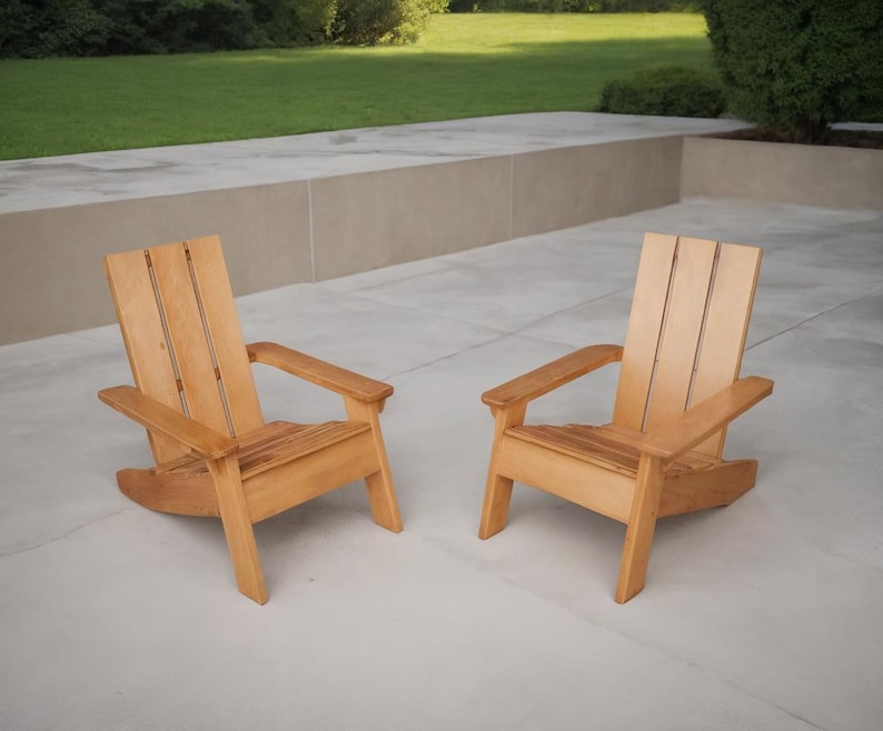 May include: Two wooden Adirondack chairs with a natural finish. The chairs have wide armrests and slatted backs. They are positioned on a concrete patio, with a grassy area and greenery in the background.
