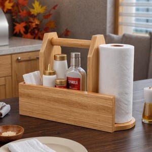 Puede incluir: Un organizador de madera de color claro con asa central, que cuenta con una varilla vertical para un rollo de papel de cocina blanco y un compartimento rectangular con una botella de vidrio transparente con etiqueta roja, dos frascos blancos y servilletas.