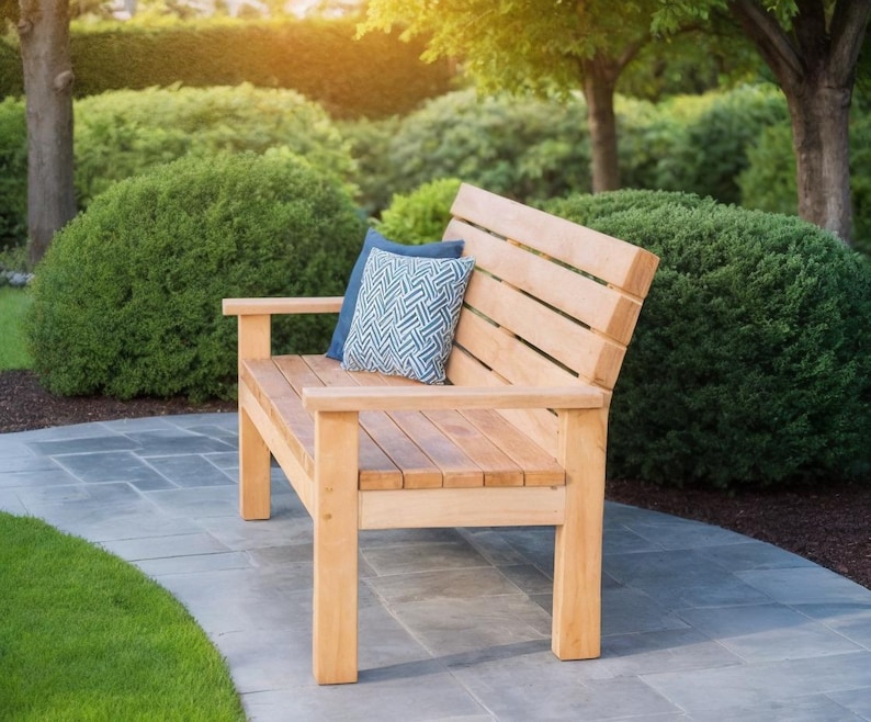 May include: A wooden outdoor bench with a slatted back and armrests. Two decorative pillows in shades of blue rest on the bench. The bench is made of light-colored wood and is set on a stone patio, surrounded by greenery.