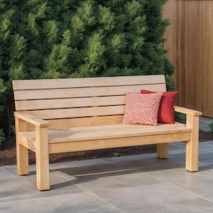 May include: A light-colored wooden bench with a slatted back and armrests. Two decorative pillows, one red and one patterned, rest on the bench. The bench is outdoors, set against a backdrop of green foliage and a wooden fence.