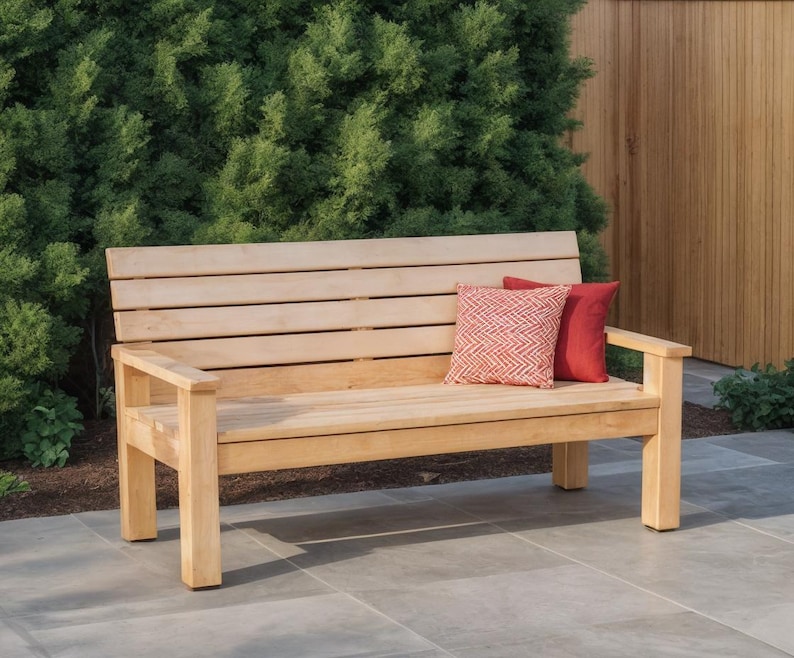 May include: A light-colored wooden bench with a slatted back and armrests. Two decorative pillows, one red and one patterned, rest on the bench. The bench is outdoors, set against a backdrop of green foliage and a wooden fence.