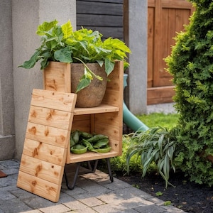 May include: A wooden plant stand with a natural finish. The stand has a rectangular shape with shelves and a hinged front panel. A large potted plant sits on top, with smaller plants on the shelves. The stand is outdoors on a brick patio.