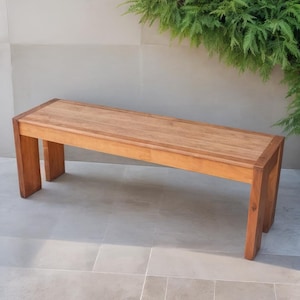 May include: A rectangular wooden bench with a light brown finish. The bench has a simple design with four sturdy legs and a flat seat. The bench is approximately 60 inches long and is placed outdoors near greenery.