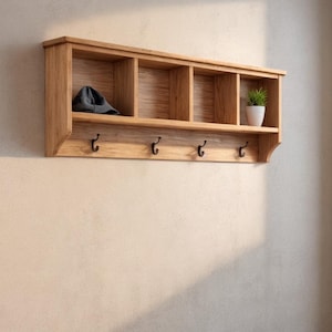 May include: A wooden wall shelf with four compartments and four hooks. A gray beanie sits in the first compartment, and a small potted plant is in the last. The shelf is mounted on a beige wall.