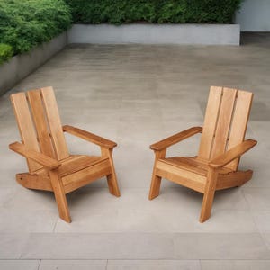 May include: Two wooden Adirondack chairs with a natural finish. The chairs have wide armrests and slatted backs. They are positioned on a light-colored stone patio, with greenery in the background.