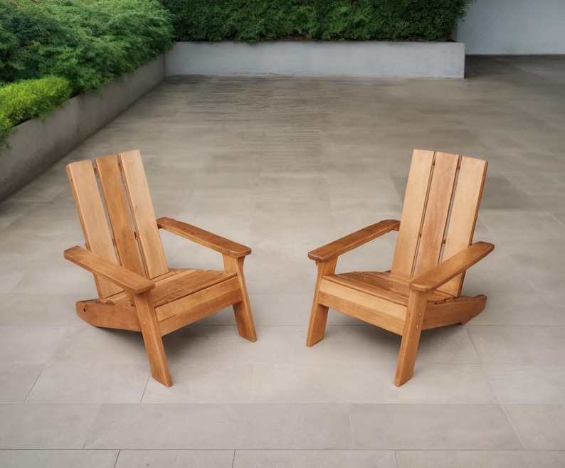 May include: Two wooden Adirondack chairs with a natural finish. The chairs have wide armrests and slatted backs. They are positioned on a light-colored stone patio, with greenery in the background.