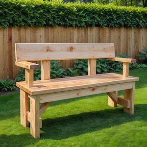 以下が含まれることがあります： 背もたれと肘掛けが付いた木製の屋外ベンチ。ベンチは明るい色の木材で作られており、緑の芝生の上に置かれています。背景には木の柵と緑の葉があります。