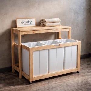 May include: A wooden laundry sorter with three white bins, a top shelf, and a sign that reads "Laundry". Two beige towels are stacked on the top shelf. The sorter is on wheels.