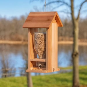 May include: A wooden bird feeder with a pitched roof and a clear plastic seed container. The feeder is filled with birdseed and is hanging from a chain. The background shows a lake and trees.