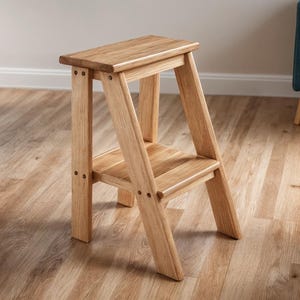 Puede incluir: Un taburete de madera clara con dos escalones. El taburete tiene un escalón superior rectangular y un escalón inferior, sostenidos por patas anguladas. La veta de la madera es visible. El taburete es de madera clara.