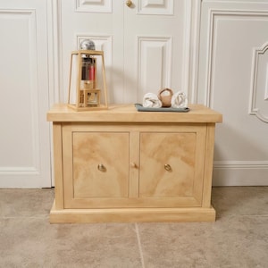 May include: A light-colored wooden cabinet with two doors and brass knobs. A decorative lamp with a glass bulb and a tray with rolled towels and a small basket sit on top. The cabinet is against a white door.
