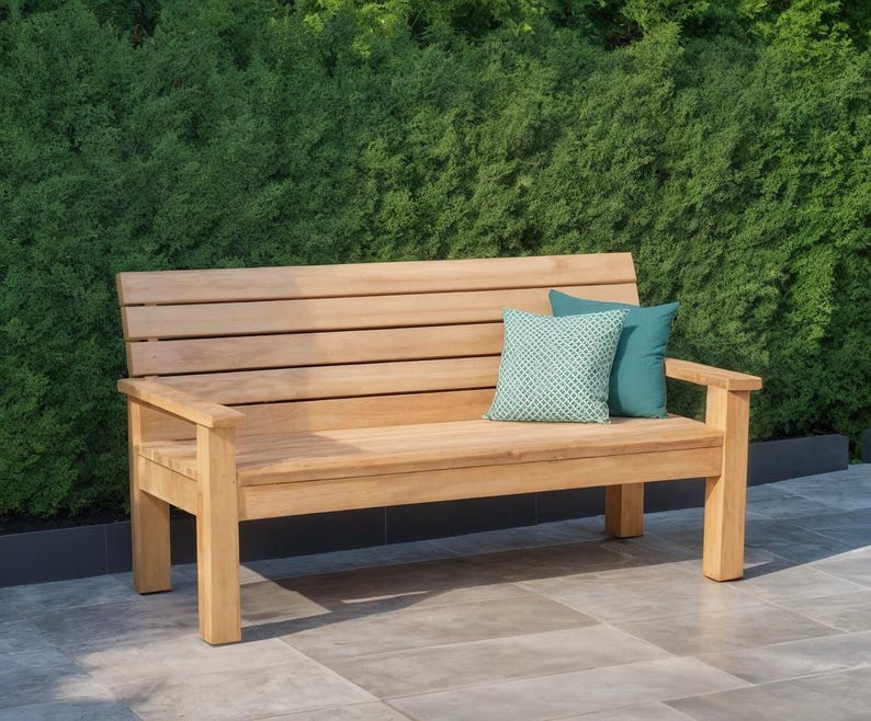 May include: A wooden outdoor bench with a slatted back and armrests. Two decorative pillows in teal and patterned green rest on the bench. The bench is made of light-colored wood and is set against a backdrop of green foliage.