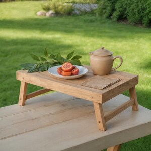 May include: A wooden breakfast tray with a teapot, a plate of sliced fruit, and a small paper placemat. The tray is set on a light-colored wooden table, with a green lawn in the background. The teapot is a light brown color.