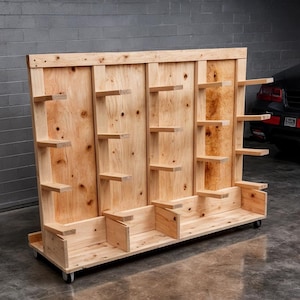 Op de afbeelding: Een grote, onafgewerkte houten displaystandaard op wielen. De standaard heeft meerdere planken en compartimenten, gemaakt van lichtgekleurd hout. Het ontwerp is eenvoudig en functioneel, geschikt voor het presenteren van diverse items. De standaard staat in een garage.