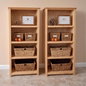 May include: Two light-colored wooden bookshelves with multiple shelves. Each shelf holds woven baskets, framed pictures, and decorative spheres. The shelves are against a neutral-colored wall and sit on a tiled floor.