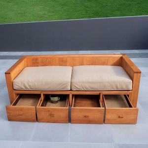 Puede incluir: Un sofá de exterior de madera marrón claro con dos cojines beige y cuatro cajones extraíbles. Una pequeña taza de cerámica se encuentra en uno de los cajones abiertos. El sofá está sobre una superficie de baldosas grises con césped verde al fondo.