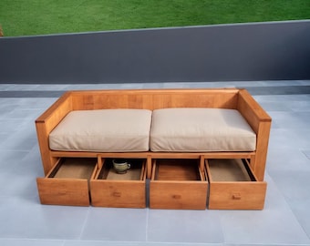 May include: A light brown wooden outdoor sofa with two beige cushions and four pull-out drawers. A small ceramic mug sits in one of the open drawers. The sofa is on a gray tiled surface with a green lawn in the background.