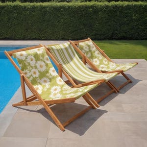 Puede incluir: Tres tumbonas de madera con asientos de lona. Dos sillas tienen un fondo verde con estampados de margaritas blancas, y una tiene rayas verdes y blancas. Las sillas están colocadas en un patio junto a una piscina.