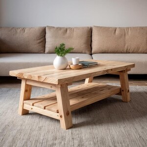 May include: A light-colored, rectangular wooden coffee table with a lower shelf. The table is placed in front of a beige sofa. A small white vase with greenery, a small wooden bowl, and a book are on the table.