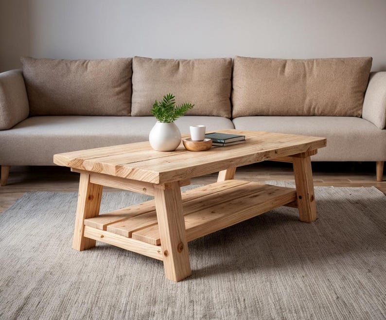 May include: A light-colored, rectangular wooden coffee table with a lower shelf. The table is placed in front of a beige sofa. A small white vase with greenery, a small wooden bowl, and a book are on the table.
