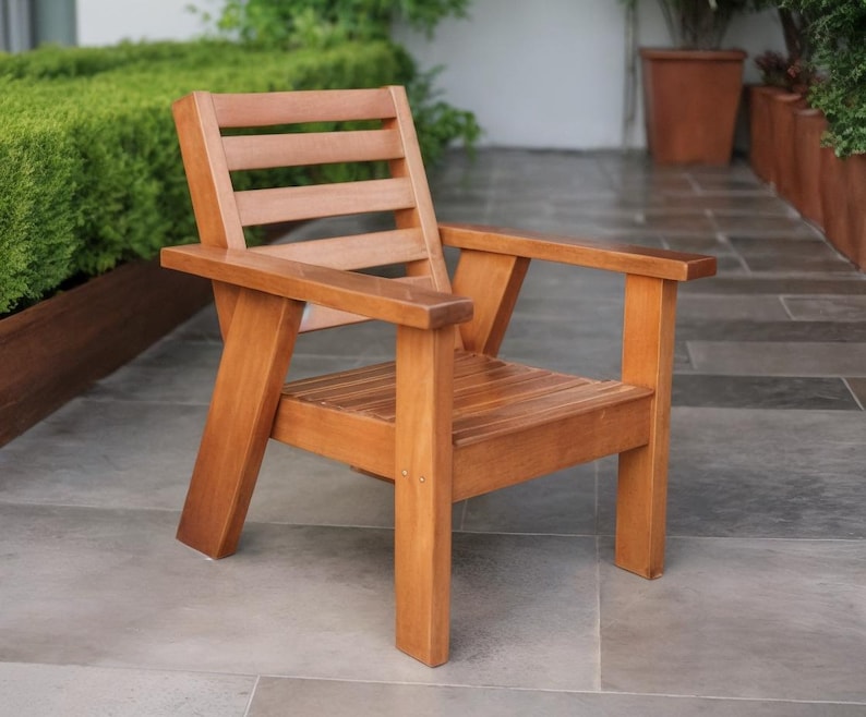 May include: A wooden outdoor chair with a natural finish. The chair has a slatted back and seat, with armrests. The chair is made of solid wood and is designed for outdoor use. The chair is approximately 61 cm wide, 61 cm deep, and 76 cm tall.