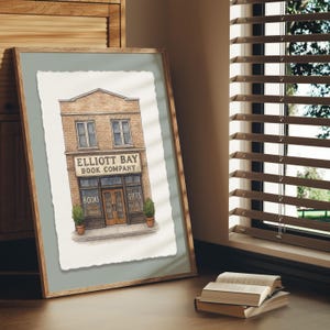 Puede incluir: Una impresión en acuarela enmarcada del edificio de la librería Elliott Bay. El edificio de ladrillo de dos pisos tiene un letrero sobre la entrada y las palabras "Books" y "Gifts" en las ventanas. La impresión se exhibe sobre una superficie de madera.