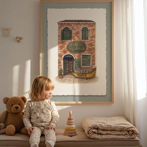 Puede incluir: Una impresión en acuarela enmarcada de una librería veneciana, "Libreria Acqua Alta", con una góndola amarilla. Un niño pequeño con un mono con estampado de margaritas está sentado en un banco con un oso de peluche, un juguete apilable de madera y una manta a cuadros.