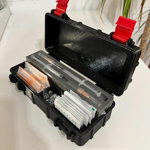 Puede incluir: Maletín negro rectangular con cierres rojos, abierto para revelar suministros médicos. En el interior hay jeringas, viales y paquetes. El maletín está sobre una superficie blanca, con una planta en el fondo.