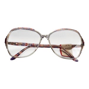May include: Vintage oversized eyeglasses with clear lenses. The frames are a light gray with floral patterned temples in shades of pink, purple, and blue. A price tag is attached to the right lens. The glasses have a retro, classic style.
