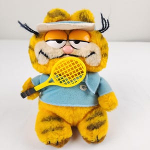 Könnte beinhalten: Ein Garfield-Plüschtier in einem blauen Tennis-Outfit und einem weißen Visier. Die orangefarbene Katze hält einen gelben Tennisschläger. Das Spielzeug hat schwarze Schnurrhaare und eine rosa Nase. Das Outfit hat ein kleines "G" auf der Brust.