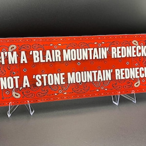 May include: A rectangular red sign with white text that reads "I'M A 'BLAIR MOUNTAIN' REDNECK NOT A 'STONE MOUNTAIN' REDNECK." The sign has a paisley pattern border and is displayed on a clear stand.
