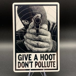 May include: Black and white sticker showing a person in a balaclava aiming a gun. The sticker includes the text "GIVE A HOOT DON'T POLLUTE" in white, bold letters. The image is displayed on a clear stand.