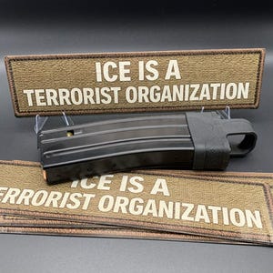 以下が含まれることがあります： 黒い銃器マガジンと、白い文字で「ICE IS A TERRORIST ORGANIZATION」と書かれた茶色のパッチ。いくつかの追加パッチが見えます。マガジンには弾薬が入っています。