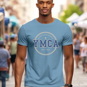 May include: A light blue t-shirt featuring the YMCA logo in a circular design. The logo includes the text "YMCA" in blue, with "Boston Massachusetts" and "EST. 1851" in a lighter color. The shirt is worn by a person.